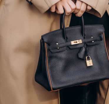 Person Holding A Small Black Handbag With A Gold Lock, Wearing A Beige Trench Coat That Whispers Hermès Secrets.