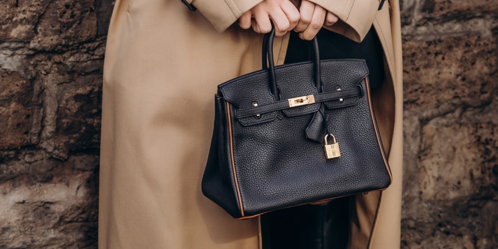 Person Holding A Small Black Handbag With A Gold Lock, Wearing A Beige Trench Coat That Whispers Hermès Secrets.