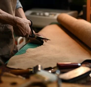 A Skilled Artisan Meticulously Cuts Semi Aniline Leather With A Knife On A Workbench, Surrounded By An Array Of Tools And Equipment.