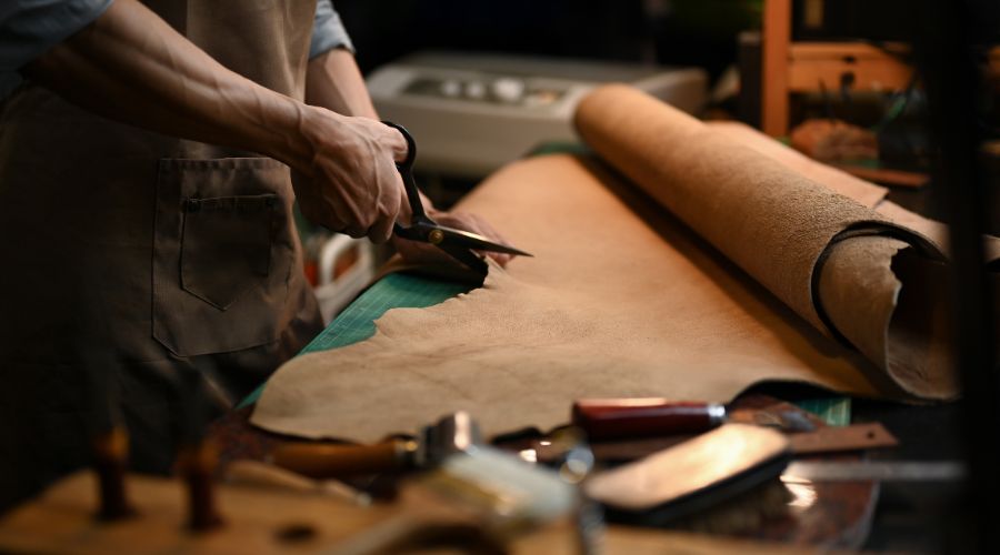 A Skilled Artisan Meticulously Cuts Semi Aniline Leather With A Knife On A Workbench, Surrounded By An Array Of Tools And Equipment.