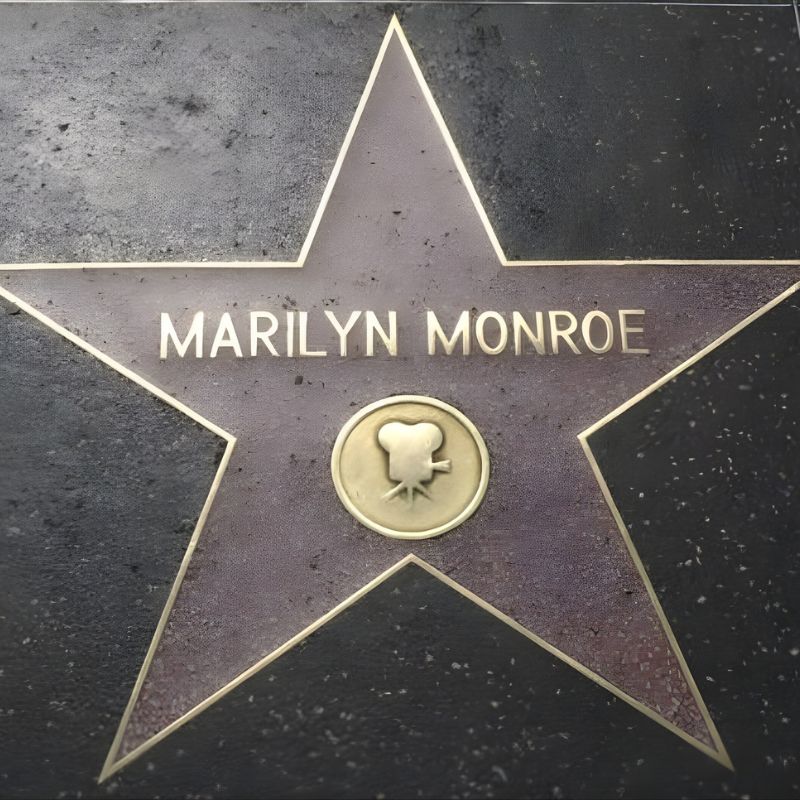 A Star Plaque On The Hollywood Walk Of Fame Displays The Name “marilyn Monroe,” Famed For Her Signature Jean Louis Designer Dress, With A Film Camera Symbol In The Center—a Nod To Her Legacy Celebrated At Many A Historic Auction.