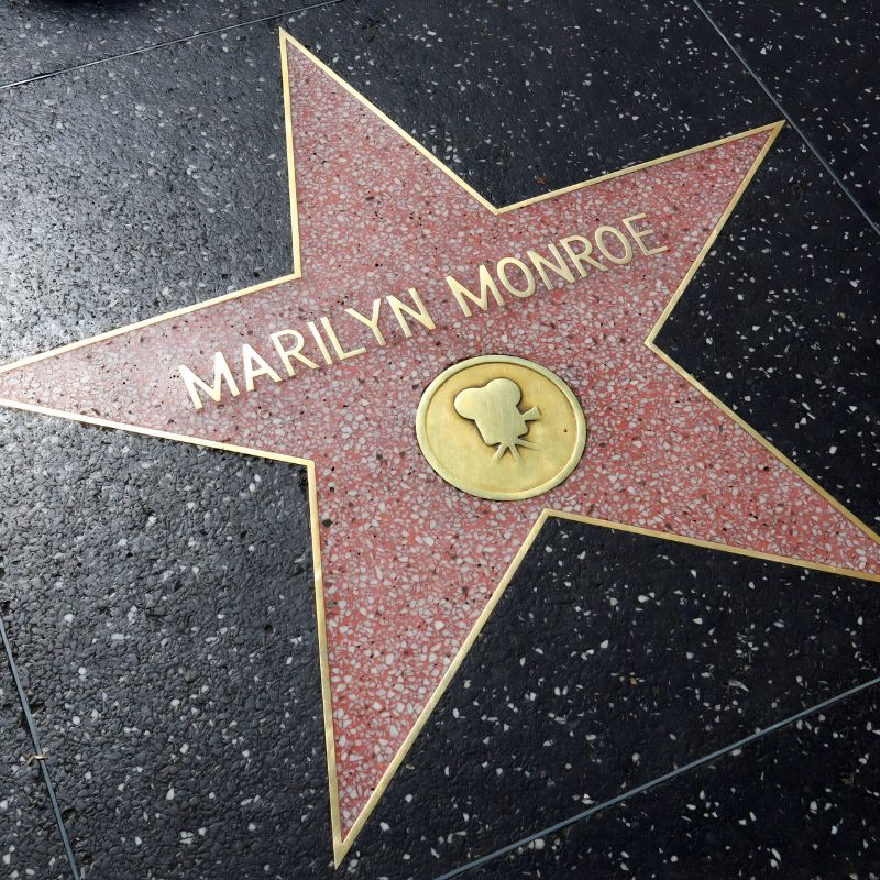 A Pink Terrazzo And Brass Hollywood Walk Of Fame Star With "marilyn Monroe" Inscribed On It, Set In A Dark Speckled Sidewalk, Celebrates Hollywood Heritage And Her Iconic Status In A Jean Louis Designer Dress.