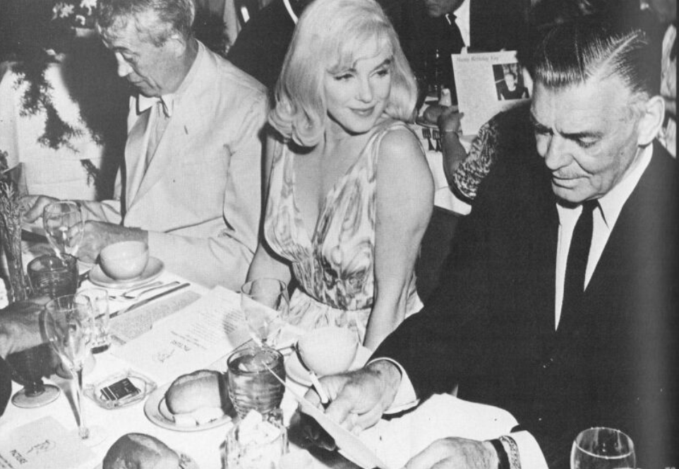 Three People Sit Together At A Formal Dinner Table, With Menus, Plates, And Glasses Visible. The Woman In The Center, Exuding Hollywood Heritage In A Jean Louis Designer Dress, Looks Toward The Man On Her Left.