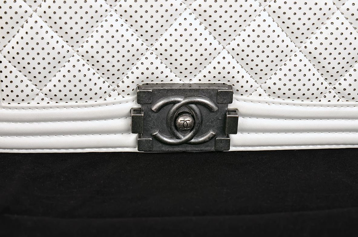 Close Up Of A Quilted White Leather Bag With Black Dots And A Metal Clasp Featuring The Chanel Logo, Positioned Above A Black Fabric Surface.