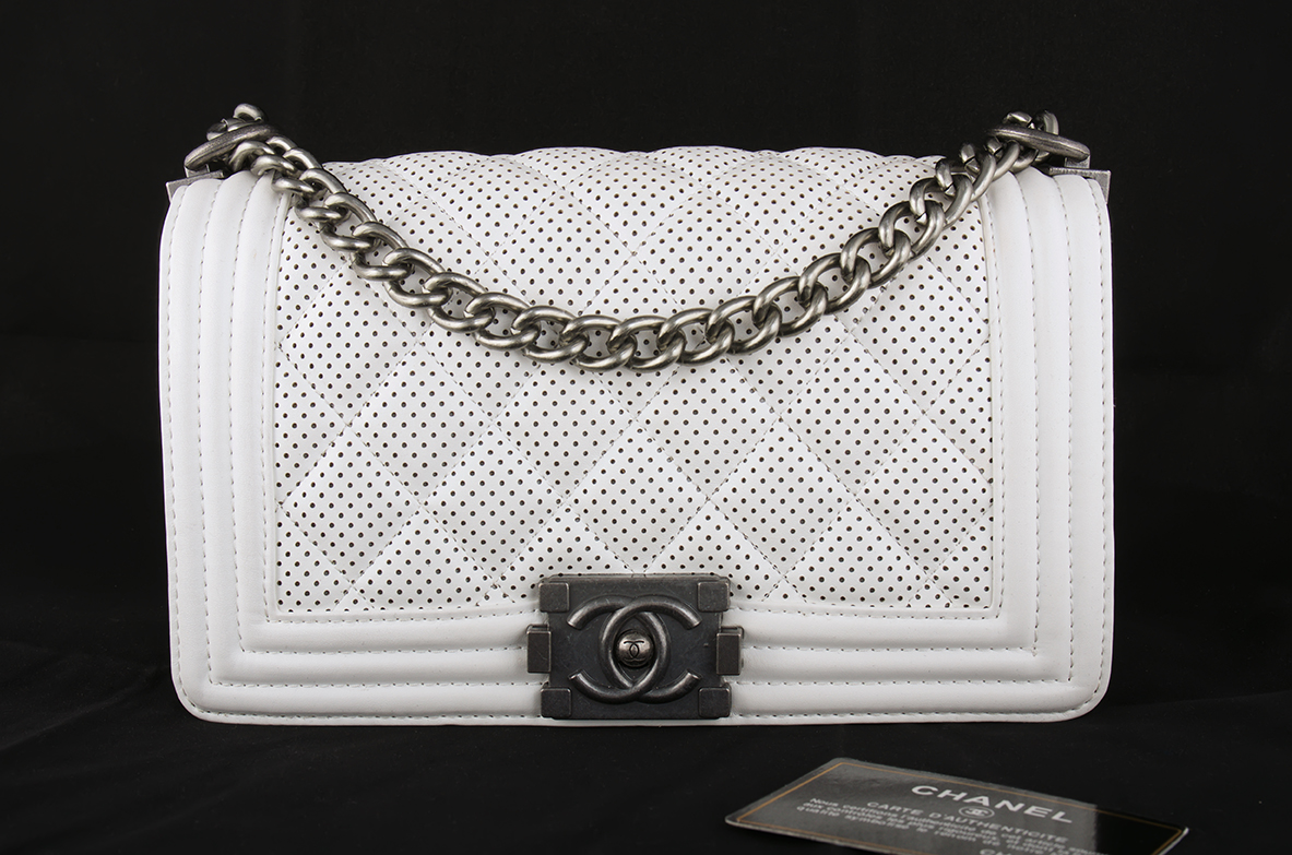 A White Chanel Handbag With Silver Chain Strap And Metallic Clasp Is Displayed Against A Black Background, With A Chanel Authenticity Card In Front.