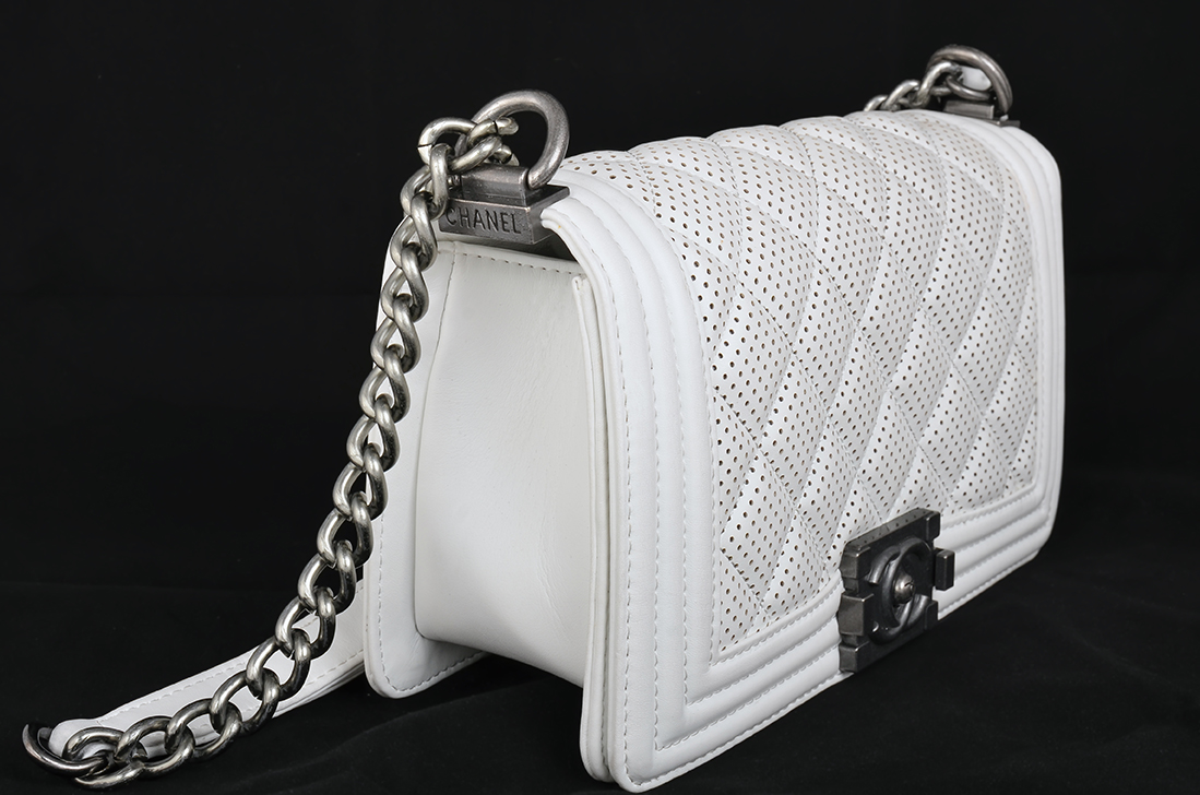 A White Quilted Leather Handbag With A Perforated Design, Silver Chain Strap, And Metal Clasp, Displayed On A Black Background.