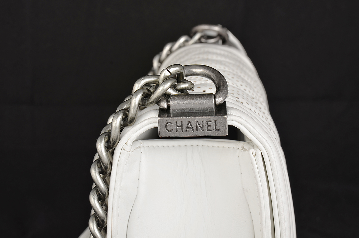 Close Up Of A White Leather Bag With A Silver Chain Strap And A Metal Clasp Engraved With The Word "chanel" Against A Black Background.