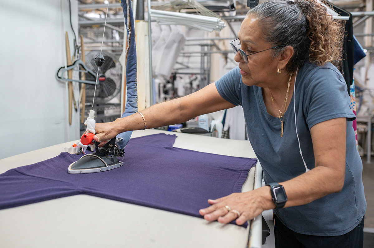 A Woman Uses An Iron To Press A Navy Blue Shirt On An Ironing Board In A Laundry Or Textile Workspace.