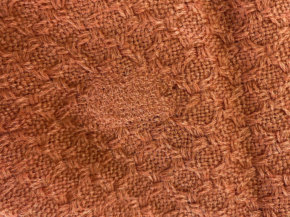 Close Up Of A Textured Orange Fabric With A Visibly Mended Patch In The Center, Showing A Different Stitching Pattern From The Surrounding Material.