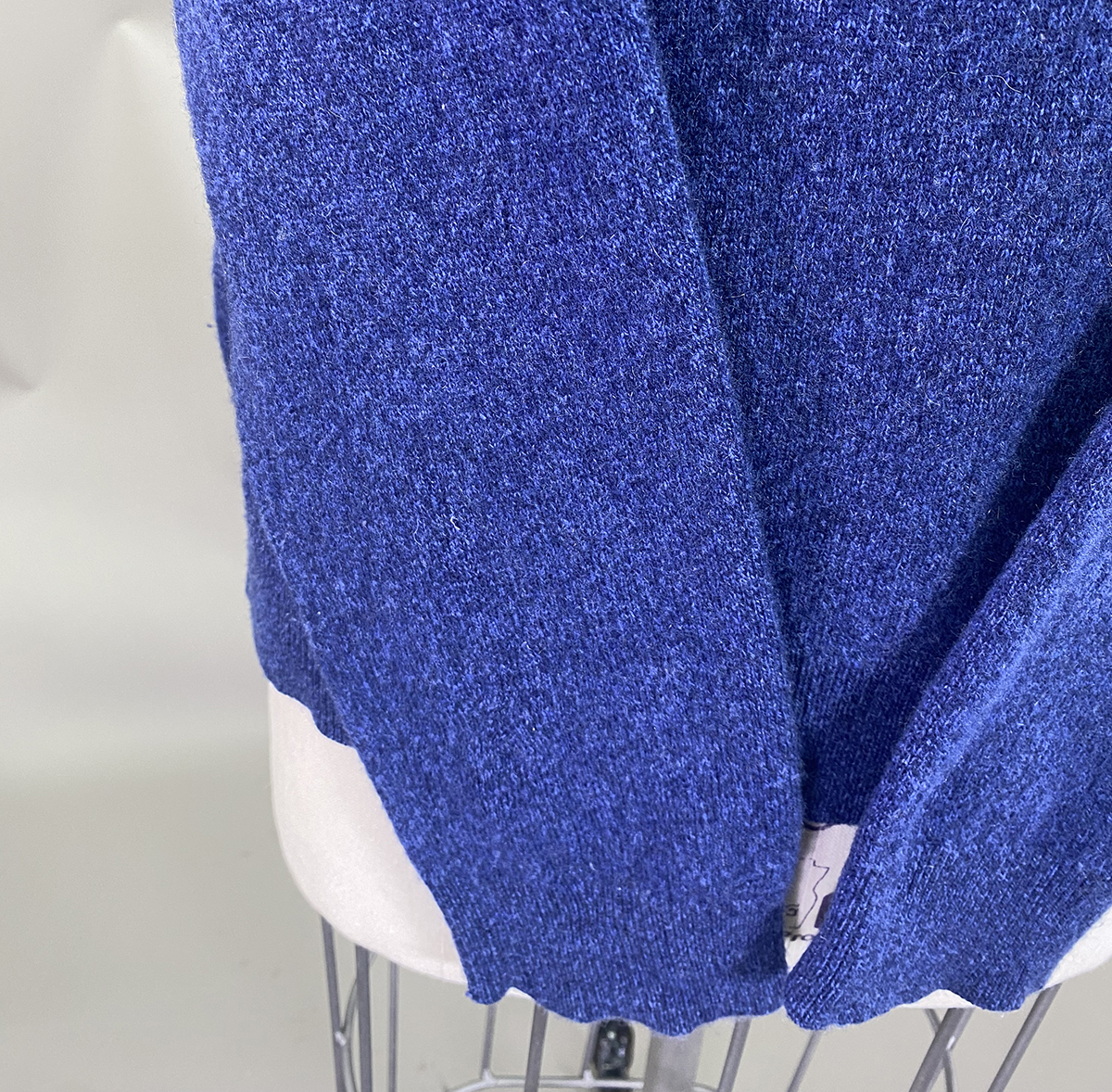 Close Up Of A Blue Knit Garment Draped Over A White Mannequin, Showing Fabric Texture And A Small Visible Clothing Tag At The Bottom Edge.