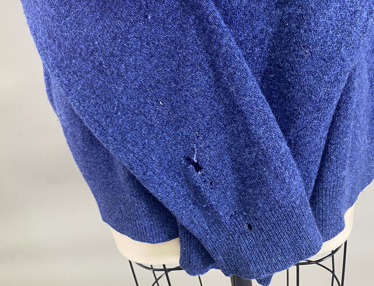 A Blue Knit Sweater With Several Small Holes And One Larger Tear Near The Bottom Hem, Displayed On A Mannequin.
