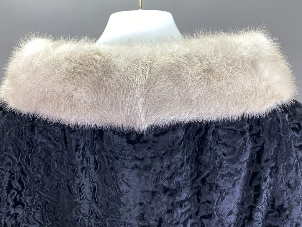 A Close Up Of A Garment On A Mannequin, Featuring Black Textured Fabric With A Light Beige Fur Collar—an Elegant Piece That Would Benefit From Expert Suede Care To Maintain Its Luxurious Look.