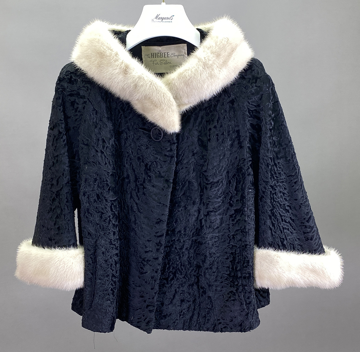 Black Textured Jacket With A Single Button, Featuring White Fur Trim On The Hood And Cuffs, Displayed On A Hanger Against A Plain Background—perfect For Those Seeking Professional Leather Care Or Tips From Our Leather And Suede Guide.