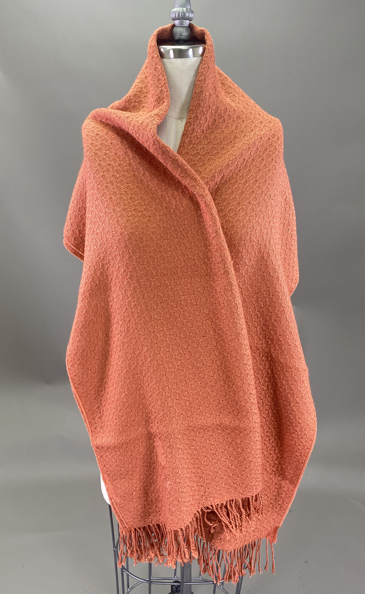 A Mannequin Draped With A Textured, Burnt Orange Woven Shawl Featuring Fringed Edges, Set Against A Plain Gray Background.