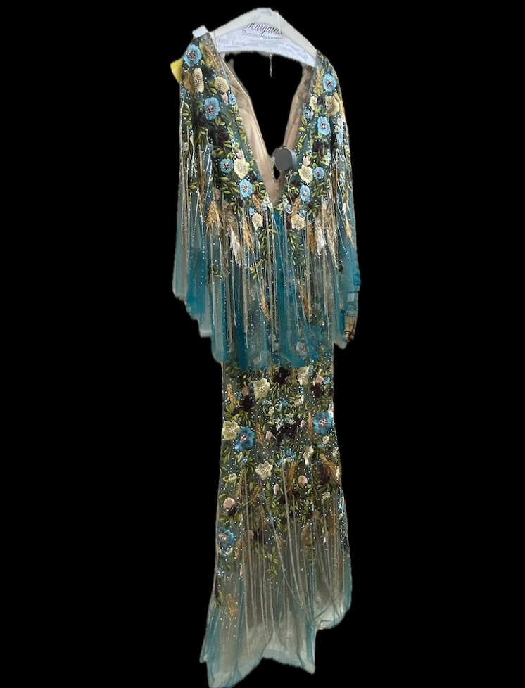 A Floor Length Gown With A Deep V Neckline, Teal Tulle Overlay, And Floral Embroidery In Blue, Gold, And Green Is Displayed On A Hanger Against A Black Background—ideal For Those Seeking Expert Gown Preservation.
