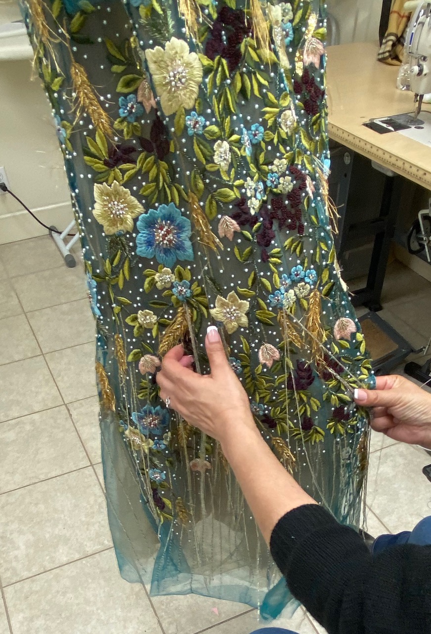 Person Holding A Sheer Fabric With Colorful Embroidered Flowers And Leaves, Next To A Sewing Machine On A Tiled Floor—perfect For Those Interested In Gown Preservation Or Beaded Gown Care.